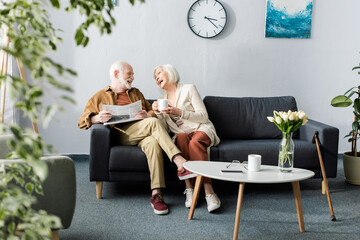 selective focus of happy senior couple laughing while talking on sofa near coffee table with tulips