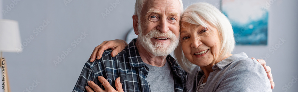 horizontal image of happy senior couple smiling and embracing while looking at camera - obrazy, fototapety, plakaty 