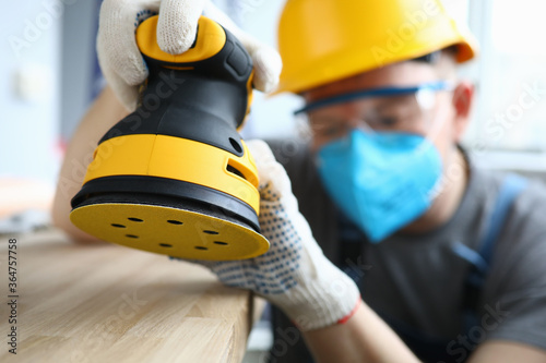 Close-up of professional worker using yellow sander equipment. Handyman polishing light wooden ground. Man wearing protective blue face mask. Renovation and construction site concept photo