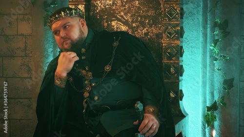warlike medieval courageous strong man sits on old bronze throne, image tyrannical king. Carnival vintage costume velvet clothes Royal crown, jewelry gems. Serious pensive face. gothic castle room  photo