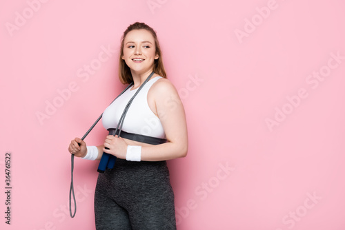 smiling overweight girl with jumping rope around neck looking away on pink