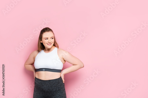 cheerful overweight girl in sportswear holding hands on hips and looking away on pink
