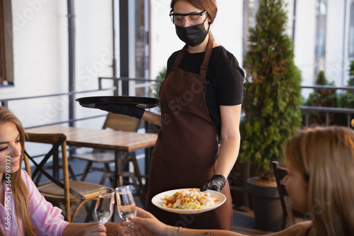 The waitress works in a restaurant in a medical mask, gloves during coronavirus pandemic. Representing new normal of service and safety. Putting the order, taking care of clients, visitors.