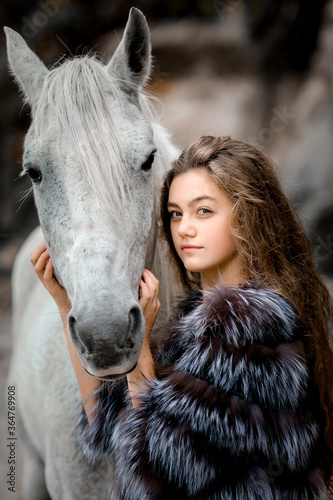 a teenage girl with long curly hair in a fur coat embraces a white horse.the open air, against the background of rocks