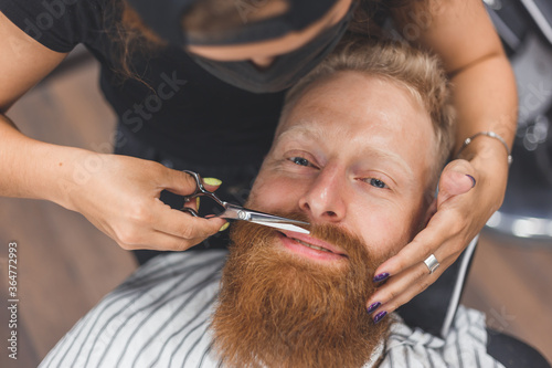 A man at a barbershop. Woman barber clipping mustache. Barber woman in mask.