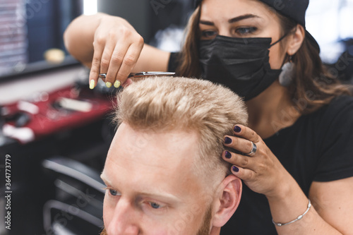 Women barber makes flat top haircut with scissors a bearded man. Service in a mask from virus.