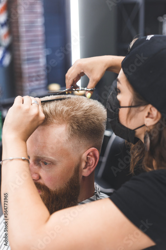 Women barber makes flat top haircut with scissors a bearded man. Service in a mask from virus.