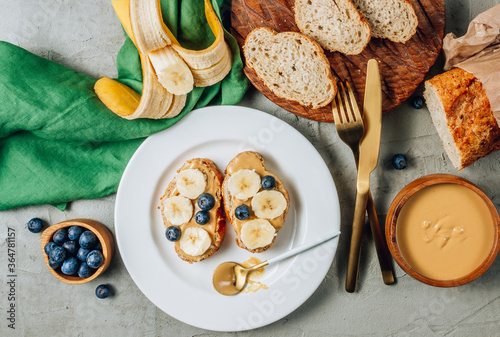 Buckwheat healthy bread with peanut butter, banana and blueberry on white plate over concrete background. Top view. Flat lay. Summer breakfast.