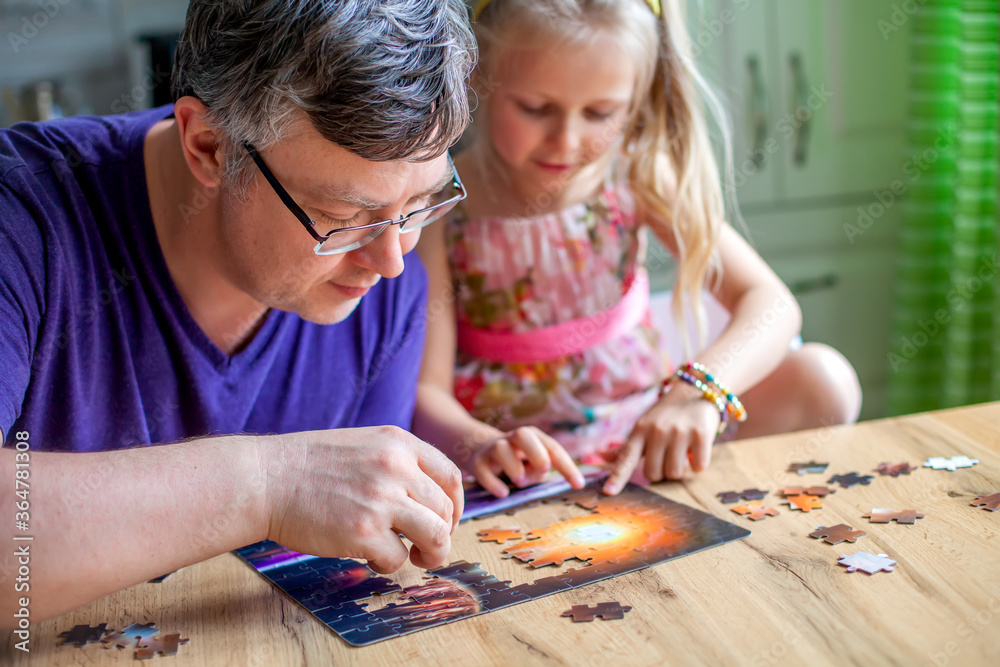 Fototapeta premium A girl sits at a table and collects a puzzle at home. He looks at the camera and laughs..Learning through the game. Learning the concentration . We play at home with parents during bad weather 