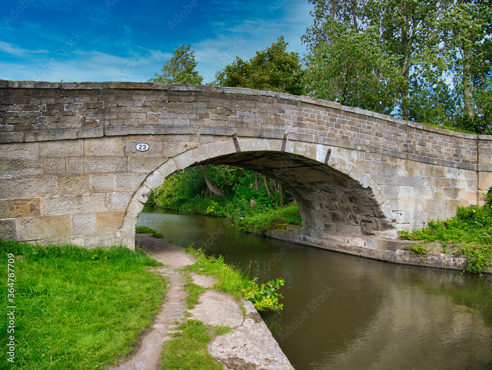 Fototapeta premium Sandstone Bridge 23 on a quiet, rural section of the Leeds to Liverpool Canal in Lancashire, UK. Taken on a sunny day with blue sky and white clouds in summer.