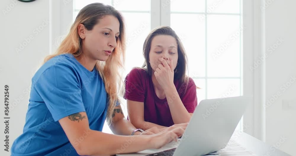 Portrait of girlfriends sitting together in the kitchen, looking at their notebook. Portrait of girlfriends sitting together in the kitchen, looking at their notebook.