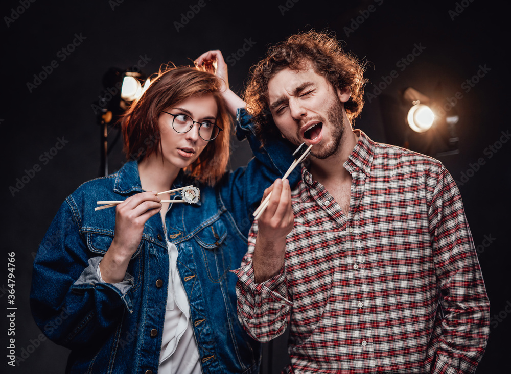 Beautiful young fashionable couple enjoys delicious sushi rolls. Dark studio with lighting equipment in the background Beautiful young fashionable couple enjoys delicious sushi rolls. Dark studio with lighting equipment in the background