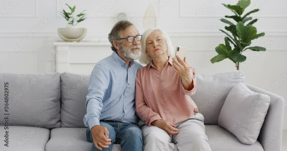 Caucasian senior man and woman woman on retirement. Happy couple sitting on sofa and posing with smiles to phone camera at home. Old couple taking selfie photos with smartphone. Caucasian senior man and woman woman on retirement. Happy couple sitting on sofa and posing with smiles to phone camera at home. Old couple taking selfie photos with smartphone.