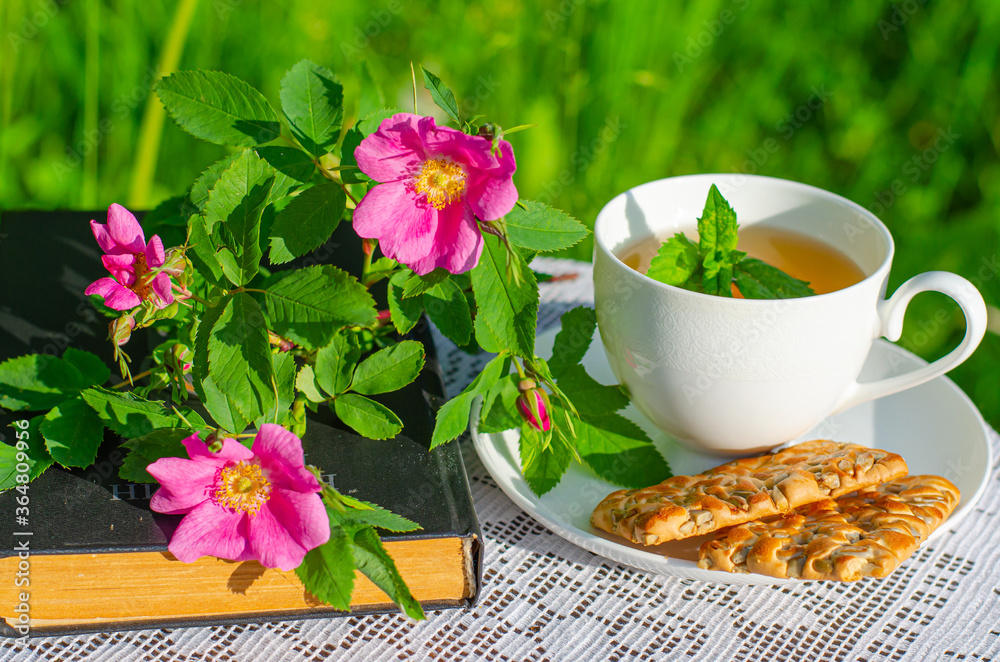 A Cup of green tea with a mint leaf, flowers, and a book on a table in the garden. Evening tea with cookies. Summer village concept. Vintage banner. A Cup of green tea with a mint leaf, flowers, and a book on a table in the garden. Evening tea with cookies. Summer village concept. Vintage banner.