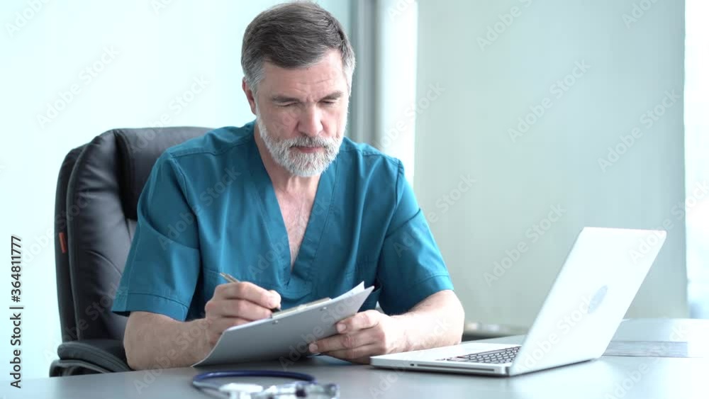 Qualified serious male therapist doing online research, reading news, making notes in medical journal. Focused old doctor physician using laptop at office desk. Qualified serious male therapist doing online research, reading news, making notes in medical journal. Focused old doctor physician using laptop at office desk.