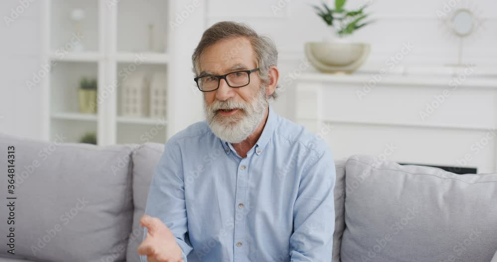 Portrait of handsome old Caucasian man blogger in glasses sitting on couch in living room and talking to camera. Male vlogger telling something and explaining. Video blogging. Vlog from home Videochat Portrait of handsome old Caucasian man blogger in glasses sitting on couch in living room and talking to camera. Male vlogger telling something and explaining. Video blogging. Vlog from home Videochat