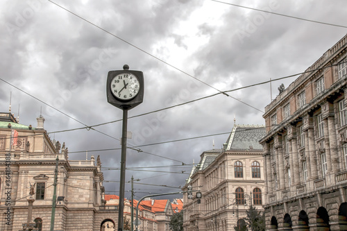 Large black metal clock with white face on metal pole in downtown Prague beneath dark clouds and overhead cables nobody