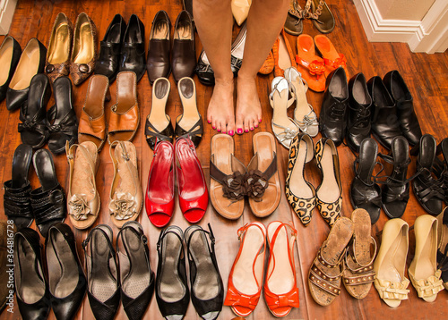 Barefoot woman’s feet surrounded by collection of shoes on wood floor