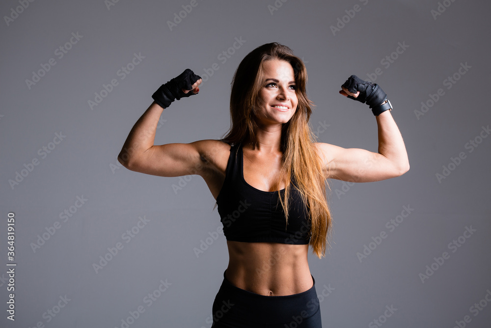 Beautiful boxer woman with long haired shows arm muscles, biceps and triceps on a gray background in gloves in a black sports suit