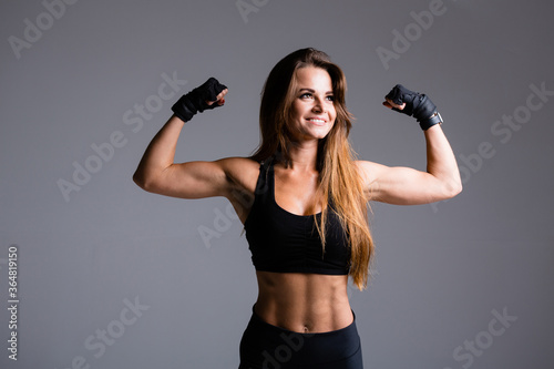 Fototapeta Naklejka Na Ścianę i Meble -  Beautiful boxer woman with long haired shows arm muscles, biceps and triceps on a gray background in gloves in a black sports suit