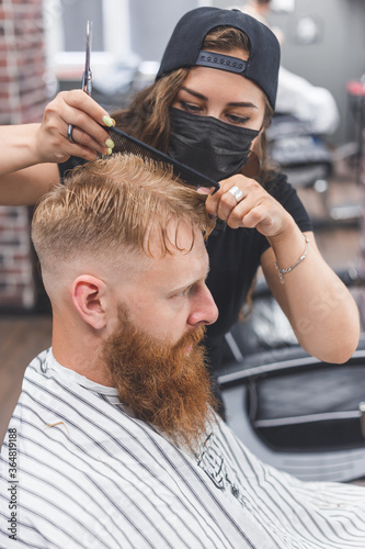 Woman barber cutting hair to a bearded man in face mask. Quarantine haircut concept.