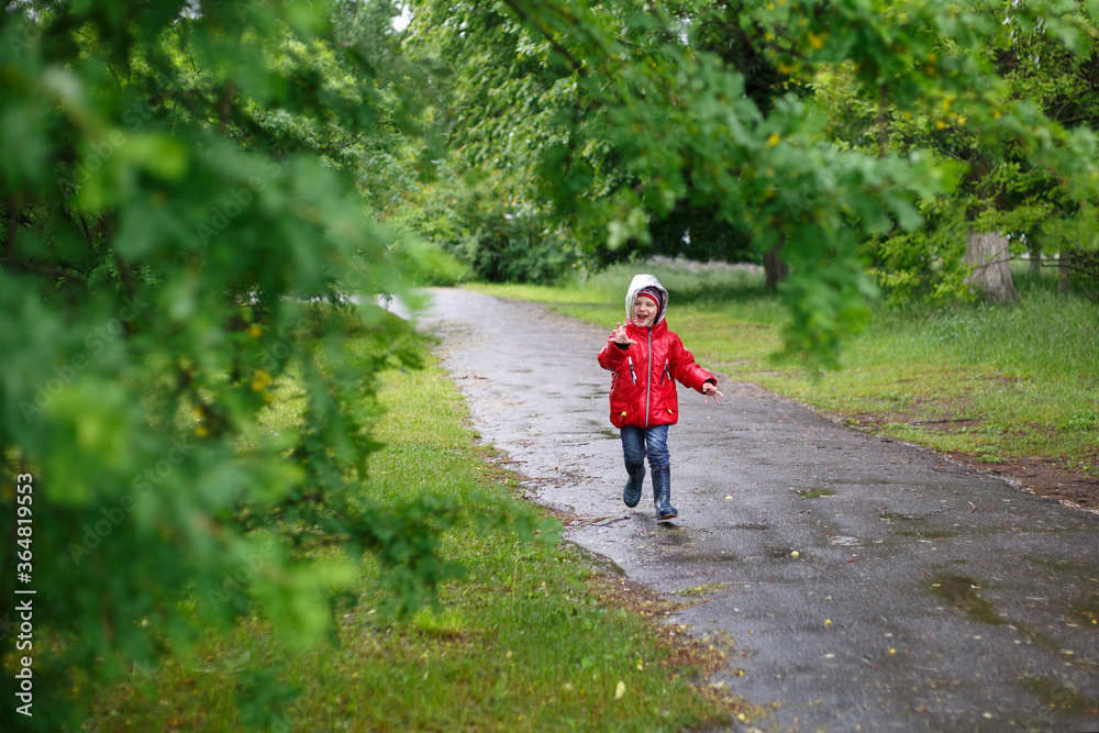 a three-year-old boy in a red jacket with a hood runs along the path from the asphalt, shows his hand and laughs a lot.
lmage with selective focus