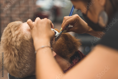 Barber woman shaves beard with a straight razor. Hairdresser equipment. Selective focus. Master in face mask