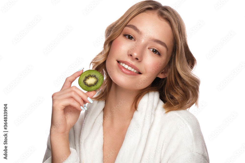 Beautiful tender young girl in a white bathrobe with clean fresh skin posing in front of the camera with kiwi in her hands. Beauty face. Skin care. Beautiful tender young girl in a white bathrobe with clean fresh skin posing in front of the camera with kiwi in her hands. Beauty face. Skin care.