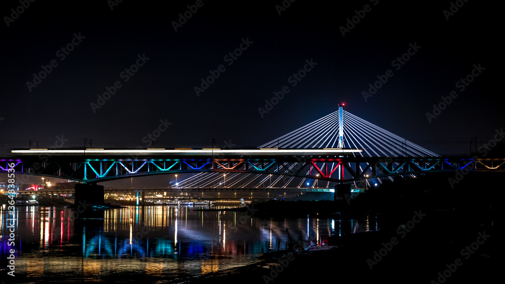 Obraz premium Night time panorama of modern european bridge. Blurred lights from passing by train. Iconic colourful bridge in Warsaw, Poland.