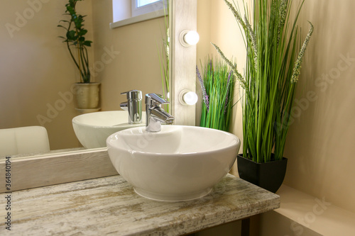 a mirror with white lights on and a white sink with two vases of green plants.