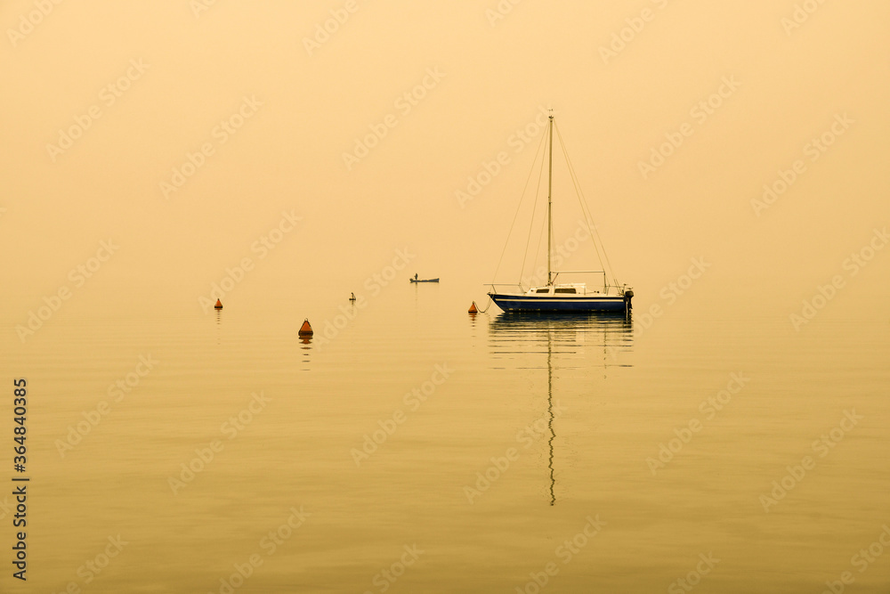 A sailboat anchored on the shore of the lake with the little boat of a fisherman on the horizon at sunset, bright sepia tone, Lake Garda, Italy A sailboat anchored on the shore of the lake with the little boat of a fisherman on the horizon at sunset, bright sepia tone, Lake Garda, Italy