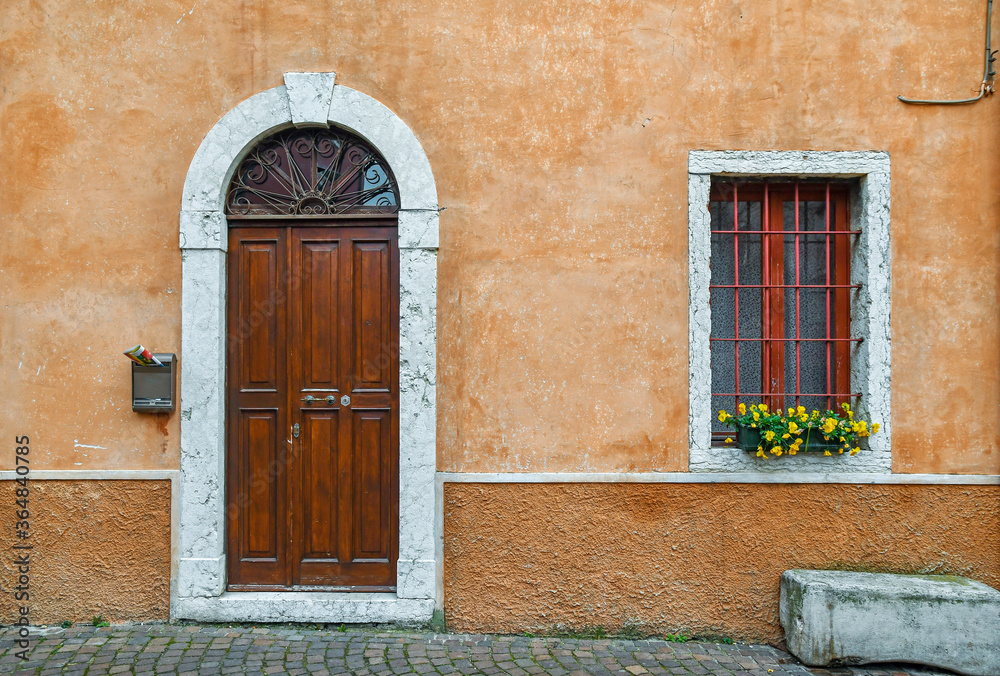 Exterior of an old building with an arched door and a window with a flowering potted plant on an orange wall, Italy