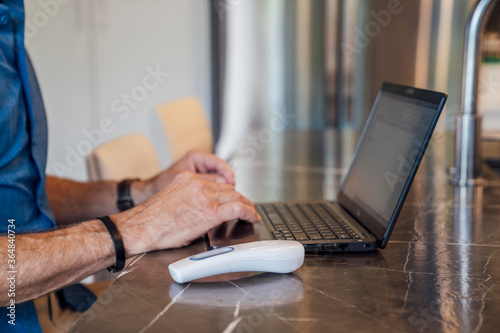 Close up of mature man's hands on keyboard of laptop with contactless thermometer next to him