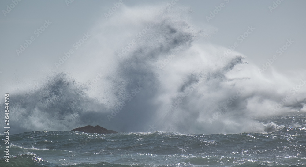 Powerful wave crashing against rocky island in ocean, with spray splashing high into the air. Misty, stormy day with high wind.