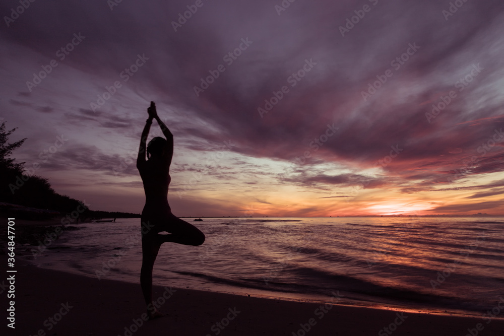 Silhouette of young woman wearing exercise clothes practicing yoga on the beach at sunset or sunrise. women do yoga. women exercising yoga in the beach. Concept : Health care