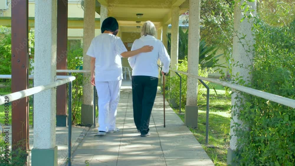 Nurse helping elderly woman with crutches at hospital. Back view