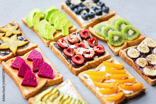 Sandwiches with cream cheese, chocolate, fresh fruits and berries avocado, blueberries, bananas, kiwi, cherry, apricot, dragon fruit on a gray concrete background. top view. Flat lay. Healthy diet