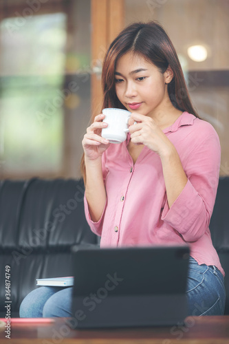 Portrait of young asian woman holding cup and looking at laptop screen while sitting in living room at home.