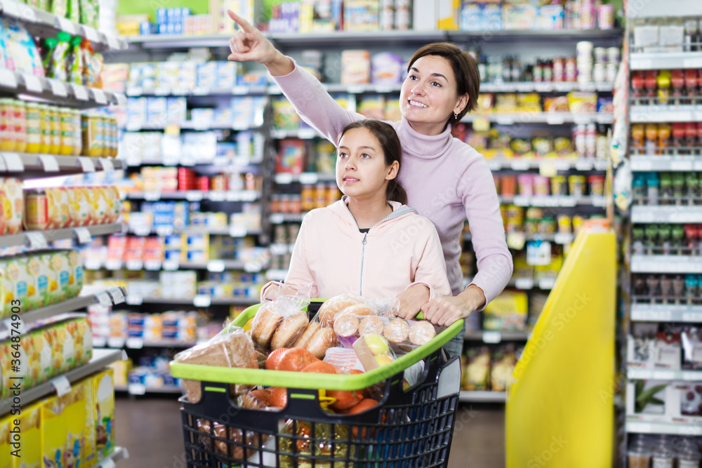 Young woman customer with girl looking for the food supplies in supermarket. Focus on both persons