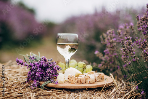 A glass of white wine, cheese, grapes, biscotti and a bouquet of flowers on a haystack among lavender bushes. Romantic picnic. Soft selective focus. photo