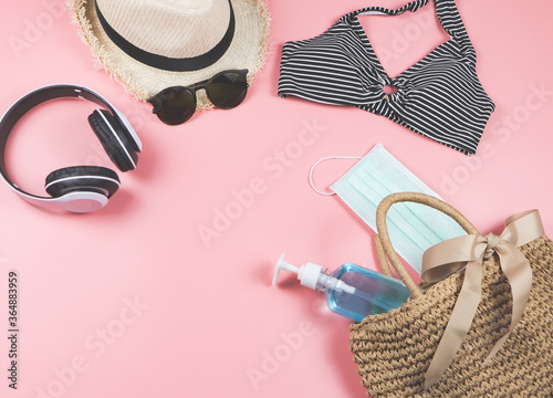 COVID-19 prevention , summer and new normal concept, top view of   bikini swim wear  and women's vacation accessories with surgical mask and alcohol  gel  in woven bag on pink background.