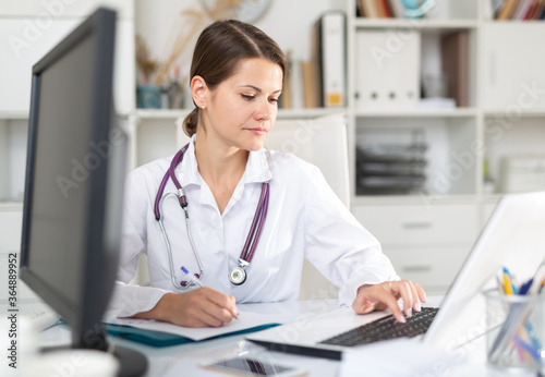 Experienced female physician filling up medical forms on laptop while sitting at table in office