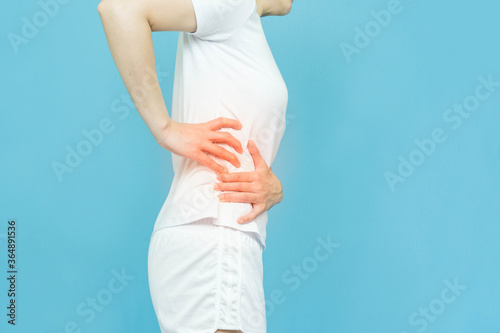 low body of human feels waist pain wearing white clothes standing over isolated blue background, hand on spot of ache and red mark, painful illness feeling unwell. health care concept. photo