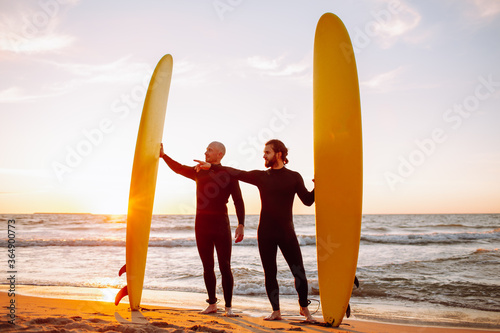 Photography Two young surfers in black wetsuit with yellow surfing longboards on a ocean coast at sunset ocean