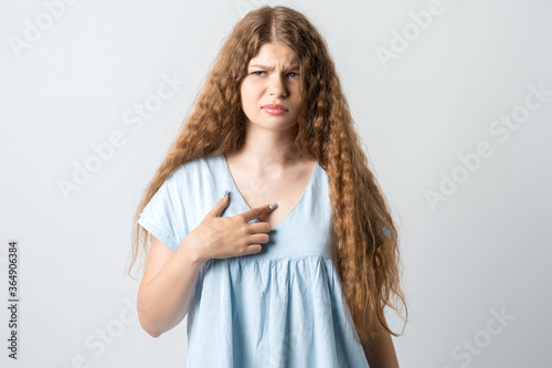Me?! Portrait of indignant girl with curly long hair startled by offensive words, points at herself with for finger, opens mouth in surprisement, has dissatisfied expression, wears casual clothes
