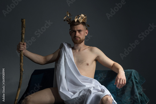 Portrait of the ancient Greek god Olympus, in white clothes, with a wreath on his head, holds a staff, sitting on a dark bench. On a gray background.