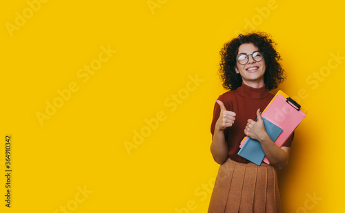 Charming brunette lady with curly hair and eyeglasses holding some folders and gesturing the like sign on a yellow studio wall with free space