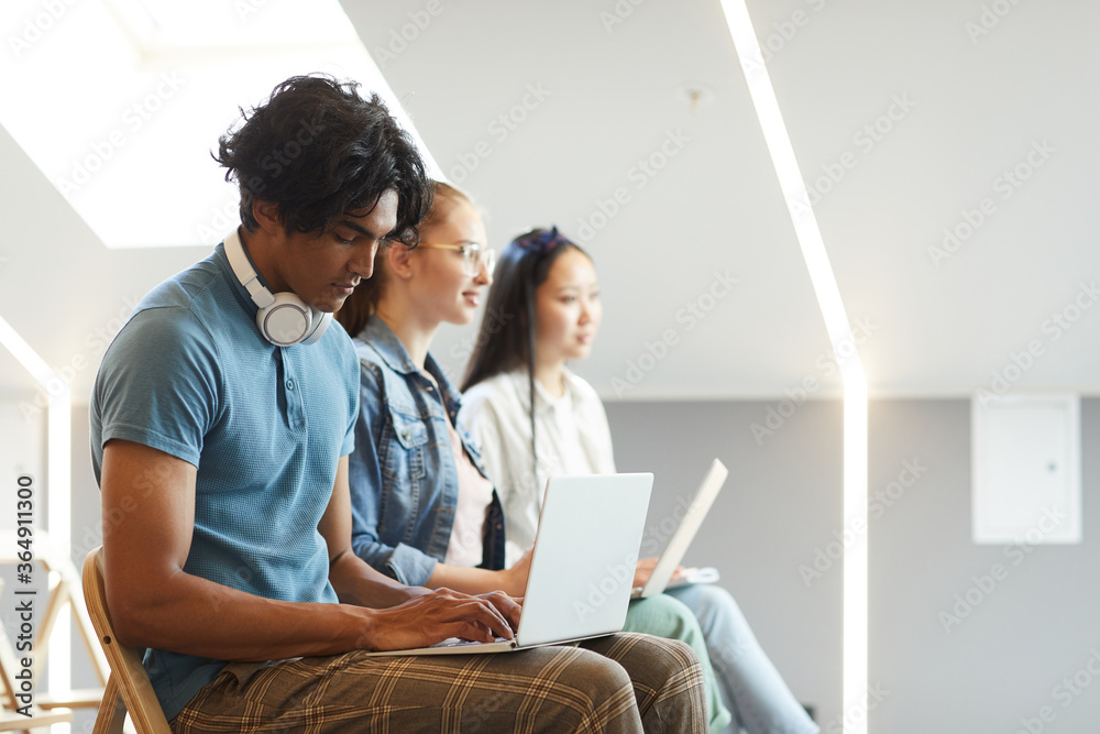 Side view of concentrated multi-ethnic students sitting in row and using laptops for notes at lecture Side view of concentrated multi-ethnic students sitting in row and using laptops for notes at lecture
