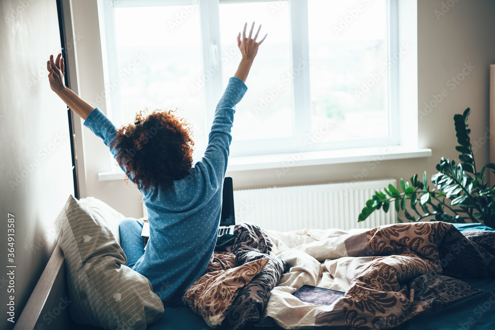 Curly haired caucasian woman in blue pajama stretching up in bed early morning - obrazy, fototapety, plakaty 