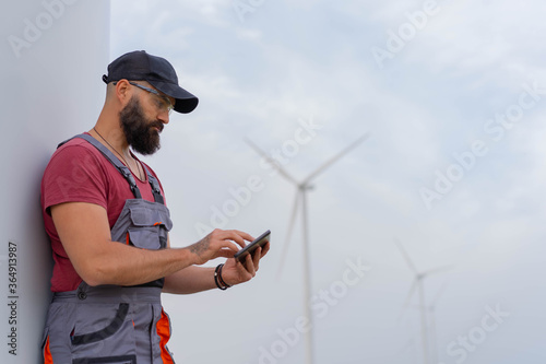 Young worker with strong beard in wind park. Working on smart phone. Wind Mill  in background.